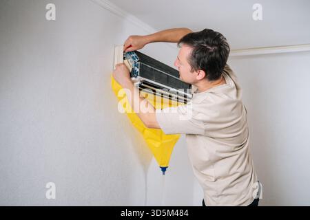 Ein erfahrener HLK-Techniker zerlegt die Klimaanlage sorgfältig und verwendet eine Schutztasche, um tropfendes Wasser und Schmutz während der Reparatur aufzufangen. Stockfoto