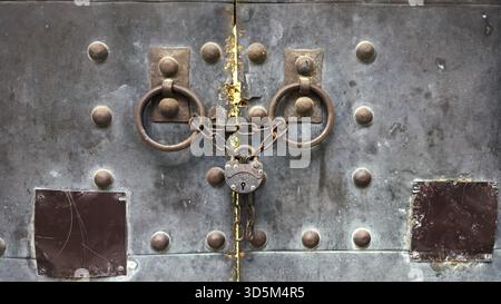 Eine Nahaufnahme einer alten Metalltür, die mit rostigen Ketten und einem alten Vorhängeschloss gesichert ist, mit schweren Eisenringen und verwitterten Texturen mit Zeichen des Alters A Stockfoto
