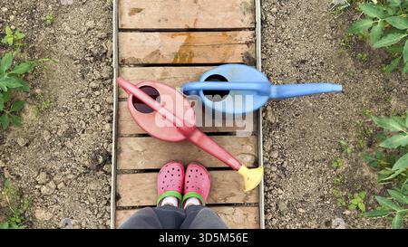 Rote und blaue Plastikgießkannen auf Holzweg im Gemüsegarten mit Person in hellen Gartenschuhen, Konzept der Gartenarbeit und Öko-Lifestyle, gard Stockfoto