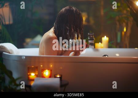 Eine Person, von hinten gesehen, mit langen, nassen, dunklen Haaren, hält es wie beim Spülen oder Auftragen der Haarmaske in einer Badewanne. Sanfte Beleuchtung durch Kerzen und umgeben von B Stockfoto
