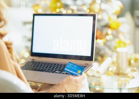 Eine Frau, die von hinten gesehen wird, hält eine Kreditkarte, während sie einen Laptop ansieht. Ein verschwommener Weihnachtsbaum mit warmen Lichtern im Hintergrund sorgt für ein festliches h Stockfoto