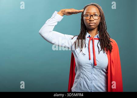 Afroamerikanerin grüßt militärisch und symbolisiert Ehre und Patriotismus. Schwarze weibliche Superheldin grüßt mit ernstem Ausdruck, Hand an Stirn, ehrt Veteranen und vermittelt Respekt. Stockfoto