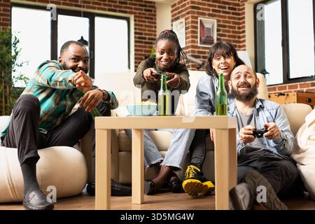 afroamerikanerin und kaukasischer Mann spielen Videospiele mit Wireless-Controllern, während Freunde beobachten und auf den Bildschirm zeigen. Verschiedene Gruppen von begeisterten Partnern spielen gemeinsam in einem modernen Wohnzimmer. Stockfoto