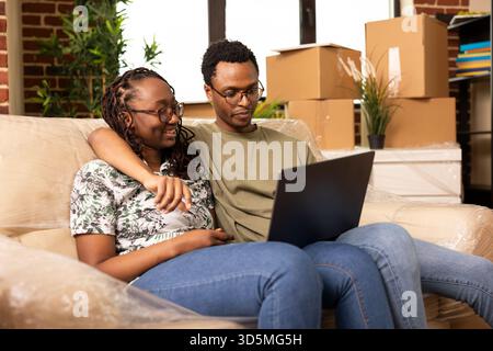 Fröhliches Paar, das auf dem Sofa mit Laptop sitzt und nach dem Einzug ein neues Zuhause plant. Afroamerikanische Partner, umgeben von ungepackten Kartons, suchen im gemütlichen Wohnzimmer online nach Inspirationen für die Dekoration. Stockfoto