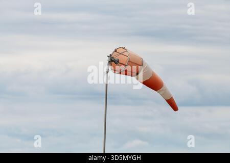 Orange-weiß gestreifte Windsocke an Metallstange, die horizontal durch Wind gegen bewölkten, grauen Himmel verlängert wird. Aufnahme auf Augenhöhe mit Indica-Windrichtung Stockfoto