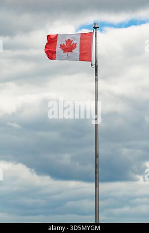 Kanadische Flagge mit rotem Ahornblatt, das auf einem hohen Metallfahnenmast vor dramatischem bewölktem Himmel fliegt. Die Flagge weht im Wind und zeigt die Bewegung an. Niedriger Winkel von Stockfoto