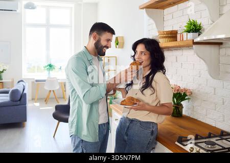 Fröhliches Paar, das Croissant und Kaffee am entspannten Morgen in der Küche teilt Stockfoto