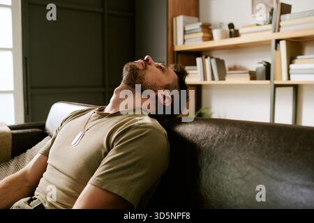 Junger Erwachsener kaukasischer Mann, der sich auf das Sofa zurücklehnt und emotional erschöpft erscheint Stockfoto