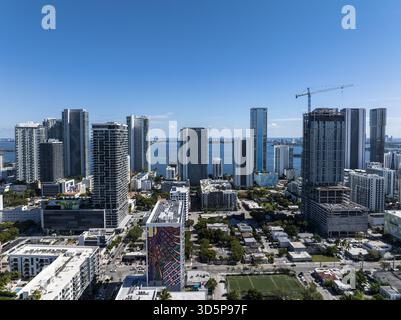 Blick aus der Vogelperspektive auf hoch aufragende Wolkenkratzer, die den hellen Himmel über der Biscayne Bay reflektieren, gegenüber den flachen Gebäuden und dem üppigen Grün der Stadt, Stockfoto