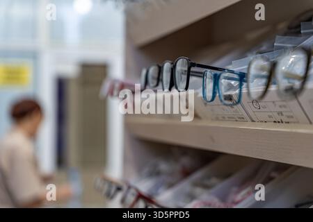 Eine Nahaufnahme der Brille, die in einem Regal in einem optischen Geschäft ausgestellt ist, mit einem unscharfen Hintergrund, der einen Kunden zeigt, der sich im Internet befindet. Stockfoto
