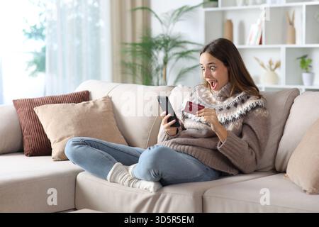 Verblüffte Frau im Winter, die online mit Kreditkarte und Kopfhörern auf einer Couch zu Hause sitzt Stockfoto