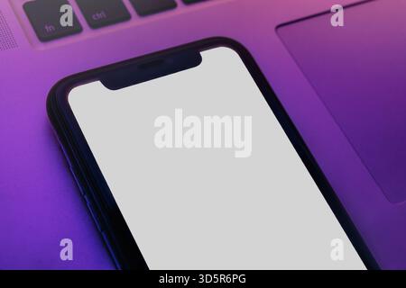 Nahaufnahme eines Smartphones mit leerem weißen Bildschirm, der auf dem Laptop unter leuchtendem rosa und lila Neonlicht liegt, Modellvorlage für die Anwendungspräsentation. Kopierbereich Stockfoto