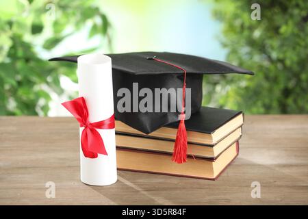 Graduierungskappe, Diplom und Bücher auf Holztisch vor verschwommenem Hintergrund, Großaufnahme Stockfoto