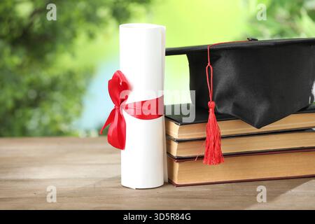 Graduierungskappe, Diplom und Bücher auf Holztisch vor verschwommenem Hintergrund, Großaufnahme Stockfoto