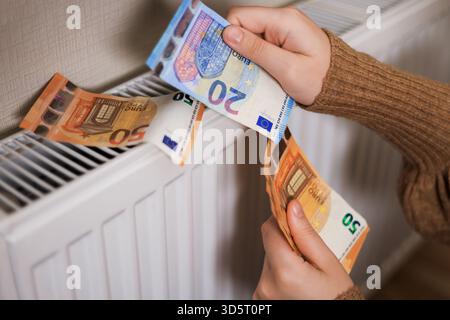 Ein Mädchen in einem Pullover zählt Geld bei einem Heizkörper: 50 und 20 Euro, eine Nahaufnahme von 20 Euro. Das Zimmer ist kalt, was den Bedarf an Heizung unterstreicht Stockfoto