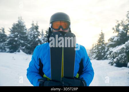 Ein Skifahrer steht in einem schneebedeckten Bereich und hält die Skier nahe am Körper. Hohe, schneebedeckte Bäume säumen den Hintergrund unter einem sanften Morgenlicht. Winter spo Stockfoto