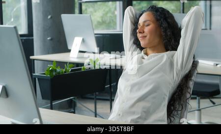 Entspannende friedliche hispanische Geschäftsfrau macht Pause im Büro Geschäftsfrau ruht Hände hinter Kopf und erledigt die Arbeit in Stockfoto