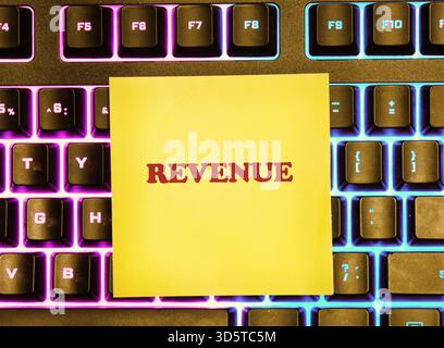 Eine leuchtend gelbe Haftnotiz mit dem Wort „Einnahmen“ wird auf einer vibrierenden, hintergrundbeleuchteten Tastatur platziert Stockfoto