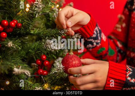 Hände legen ein rotes Glitzerschmuck auf einen Weihnachtsbaum, der mit Beeren, Lichtern und mattierten Zweigen verziert ist Stockfoto