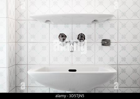 Ein sauberes und modernes, weißes, an der Wand montiertes Waschbecken aus Keramik mit Chromarmatur und schwarzen, runden Griffen. Weiße Fliesen bedecken die Wand im Hintergrund, Stockfoto