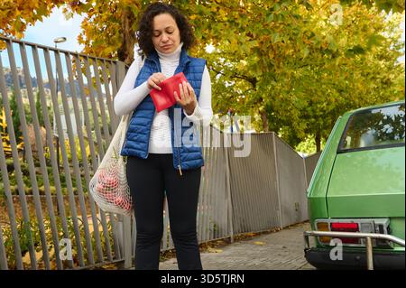 Eine Frau im Freien im Herbst untersucht eine rote Brieftasche, während sie eine blaue Puffweste und einen weißen Pullover trägt. Sie trägt einen Lebensmittelbeutel neben einem grünen Ca Stockfoto