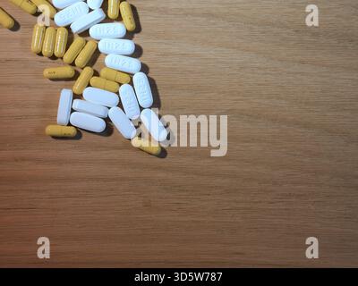 Pillen und Kapseln auf Holztisch - Weiße ovale Pillen und gelbe Kapseln verstreut auf einer hellbraunen Holzoberfläche. Symbolisiert Gesundheit, Medizin, Tre Stockfoto