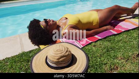 Liegende Frau in gelbem Badeanzug und Sonnenbrille am Pool mit gestreiftem Handtuch und Strohhut Stockfoto