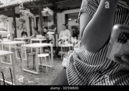Schwarz-weiß-Atmosphäre Szene eines lokalen städtischen Cafés. Eine einsame junge Frau sitzt einfach und beschäftigt mit ihrem Smartphone, während sie im Hintergrund ist, irgendeine andere Stockfoto
