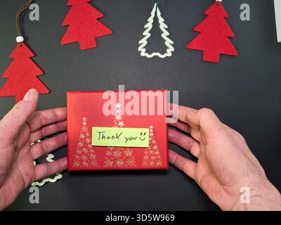 Dankeschön Geschenkbox mit Weihnachtsdekor - Überkopfansicht der Hände, die eine rote Weihnachtsgeschenkbox mit goldenen Weihnachtsbäumen und einer Handschrift präsentieren Stockfoto