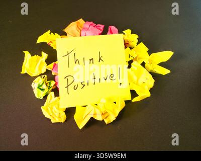 Denken Sie Positiv! Motivationshinweis – „Positiv Denken!“ Nachricht auf einem hellgelben Ton, kontrastiert mit bunten zerknittertem Papier auf dunklem Hintergrund. Symbol Stockfoto