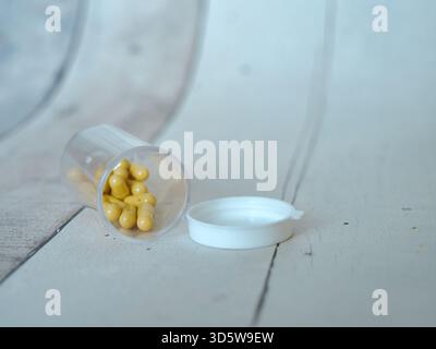 Gelbe Kapseln aus Pillenflasche verschütten - gelbe ovale Kapseln verschütten sich aus einer transparenten Arzneimittelflasche aus Kunststoff neben der weißen Kappe auf einer sauberen, lila Stockfoto