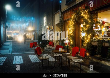 Eine ruhige Kopfsteinpflasterstraße und eine Bar-Terrasse werden an einem kühlen, nebeligen Abend in Asti, Italien, von Weihnachtslichtern und Dekorationen beleuchtet. Stockfoto