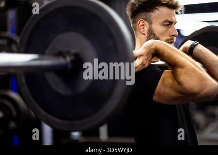 Ein Mann führt eine Gewichtheben-Übung in einem modernen Fitnessstudio aus. Er konzentriert sich intensiv, während er eine Langhantel hebt und dabei Stärke und Entschlossenheit demonstriert Stockfoto