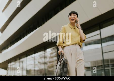 Eine fröhliche Frau in einer gelben Bluse und einer leichten Hose spaziert in der Nähe eines eleganten modernen Gebäudes. Sie führt ein Telefongespräch und strahlt ein positives vib aus Stockfoto