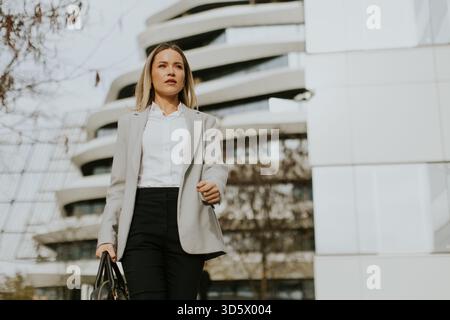 Eine Geschäftsfrau in formeller Kleidung läuft gezielt vor einem modernen Gebäude. Sie trägt eine Tasche und scheint auf ihr Ziel konzentriert zu sein. It Stockfoto