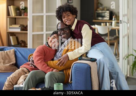 Reifer Schwarzer Mann umarmt zwei Schwarze Jungs lächelnd auf Wohnzimmer Sofa Stockfoto
