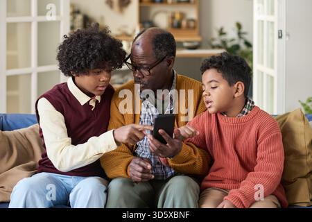 Reifer Schwarzer Mann sitzt mit zwei Jungen und benutzt das Smartphone zusammen Stockfoto