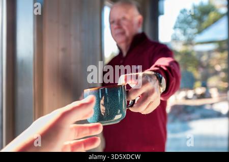 Gemeinsame Morgenbetreuung als älterer Mann bietet einer anderen Person im sanften nordischen Licht einen warmen Becher, der eine sanfte Geste der Verbindung, Freundlichkeit und s einfängt Stockfoto
