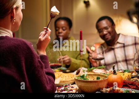 Verschiedene Gruppen von jungen Erwachsenen und Freunden mittleren Alters, die Thanksgiving-Dinner teilen, kaukasische Frau mit Gabel und Dessert im Vordergrund, Schwarzer Mann und Frau, die zusammen essen Stockfoto