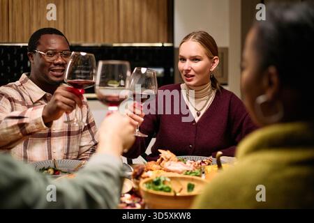 Verschiedene Gruppen junger Erwachsener und Frauen mittleren Alters, die während des Thanksgiving-Dinners unter Freunden Weingläser hochheben, lächeln und Toast am festlichen Tisch mit Essen machen Stockfoto