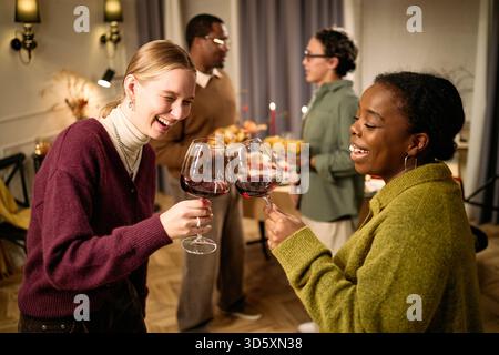 Junge Erwachsene kaukasische Frau und junge Erwachsene Schwarze Frau, die Weingläser klirrt und lächelnd, multiethnische Gruppe junger Erwachsener im Hintergrund, die während des Thanksgiving-Dinners unter Freunden Essen servieren Stockfoto