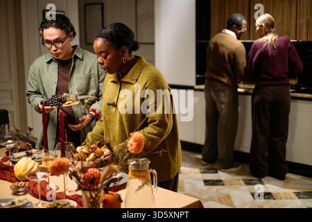 Junge Erwachsene Schwarze Frau serviert Essen am Tisch mit einem jungen Erwachsenen kaukasischen Mann, der einen Teller mit Trauben hält, multiethnische Freunde bereiten gemeinsam ein lustiges Thanksgiving-Abendessen in der Küche vor Stockfoto