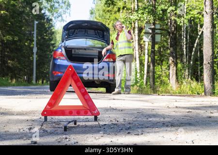 Nahaufnahme eines tragbaren reflektierenden roten Warnschilds an der Seite einer Landstraße in der Nähe eines Autos. Der Fahrer am Telefon ruft technische AS an Stockfoto