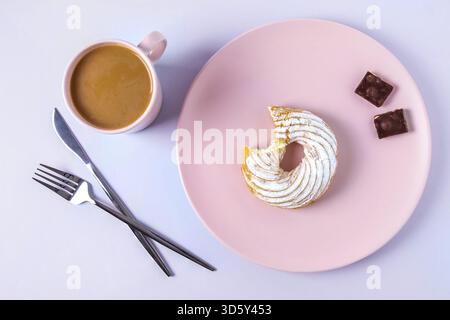 Blick von oben auf das Stillleben eines gebissenen Kuchens auf einem rosafarbenen Teller, Besteck und eine Tasse Kakao oder Kaffee mit Milch. Selektiver Fokus, horizontale Ausrichtung Stockfoto