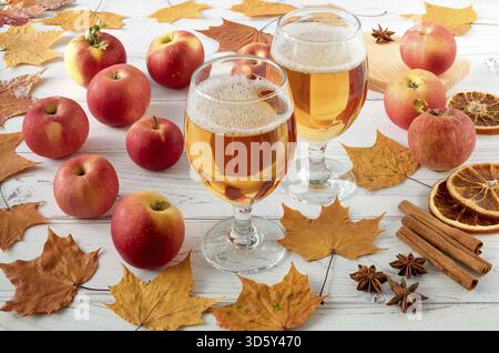 Reife frische saftige rote Äpfel, Gewürze und Gläser mit Cidre auf einer hellen Holzoberfläche mit Herbstblättern. Horizontale Ausrichtung, selektiver Fokus Stockfoto
