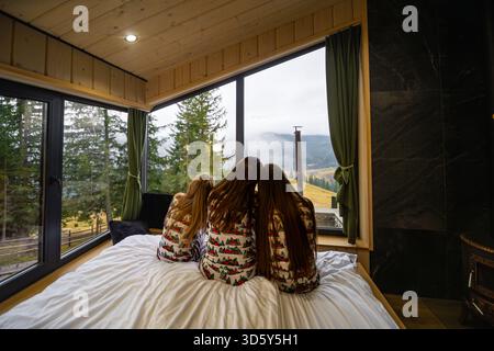 Familie im passenden Pyjama mit Blick auf die Berge. Drei Mädchen im festlichen Weihnachts-Pyjama sitzen auf einem Bett und schauen friedlich aus der großen Hütte Stockfoto
