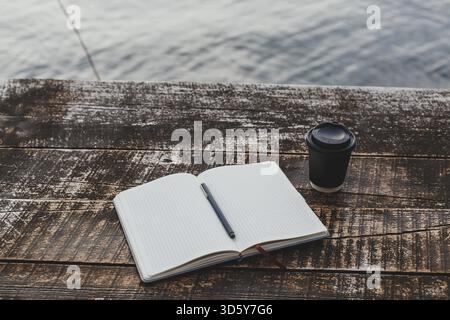 Ein Raster-Notizbuch mit Stift liegt offen auf einem rustikalen Holztisch am Wasser, neben einer schwarzen Kaffeetasse zum Mitnehmen, eine friedliche Umgebung zum Schreiben oder Refl Stockfoto