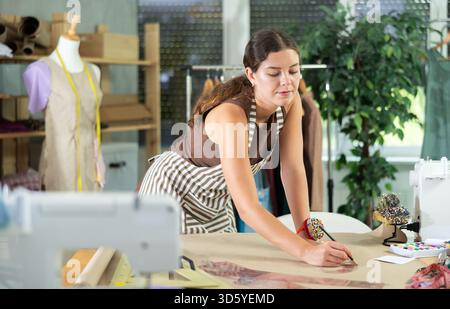 Die Schneiderin zeichnet in der Nähwerkstatt anhand von Mustern eine Skizze der zukünftigen Kleidung auf Papier Stockfoto