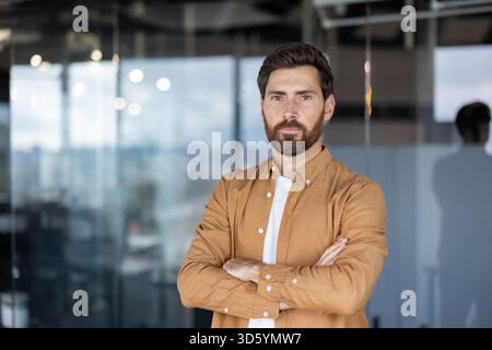 Junger bärtiger Mann posiert mit gefalteten Armen, der Selbstvertrauen und Professionalität ausdrückt, während er in einem modernen Büro steht und Führung und Erfolg repräsentiert Stockfoto