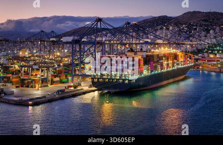Ein beleuchtetes Container-Superfrachtschiff wird abends von Kränen und Lkws in einem gewerblichen Terminal entladen und beladen Stockfoto